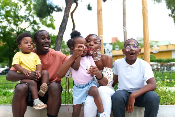 Vibrant photo: African family in a city park, parents and three kids sitting, while the girl happily blows bubbles, spreading joy.