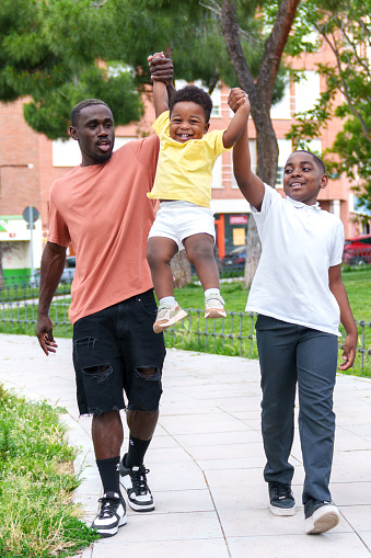 A vibrant, vertical photo captures an African family's exuberance as a young father and two sons gleefully leap high, hand in hand, in a colorful city park.