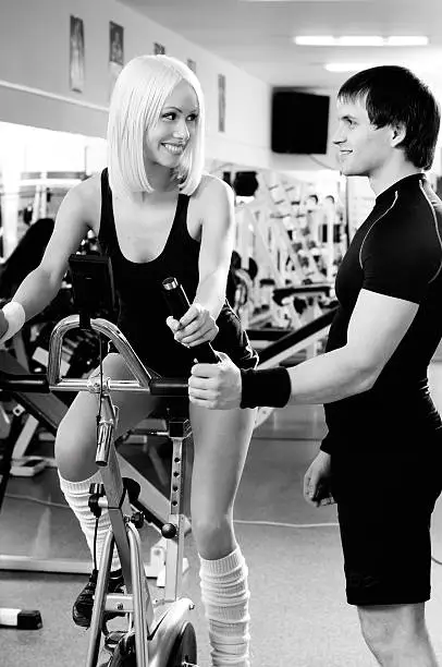 guy instructor,  give instructions cutie girl, about execute exercise on exercise-bicycle and smile, in  sport-hall, blak and white photo