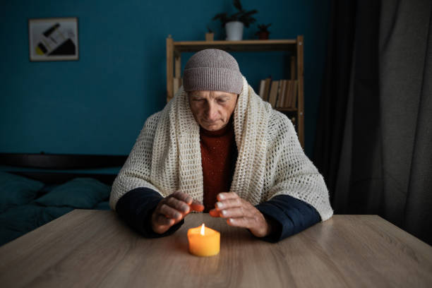 istock Old man dressed in a warm winter clothes sits at home at a table and warms his hands from a burning candle. Concept of no heating in winter at home. 1542305817