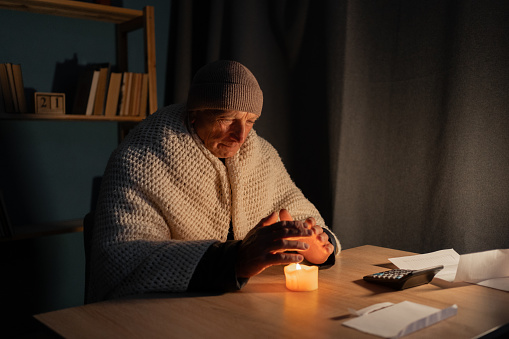 Freezing old man in winter clothes warms his hands over candle as energy blackouts cause electricity outages. Copy space