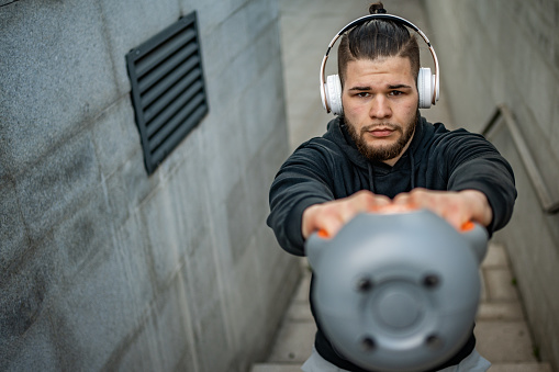 A young athlete exercises with kettlebells outdoors, dressed in sportswear, standing on the steps in front of a concrete wall