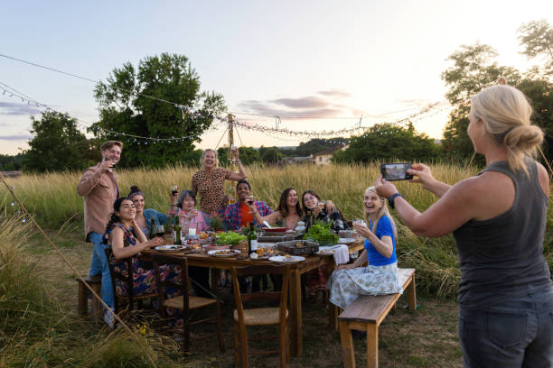 Smile Please, Say Cheers. An elevated image of a group of friends sitting at a laden dining table out in a field on a summers evening, they are toasting the camera held by one of the woman. big family table stock pictures, royalty-free photos & images