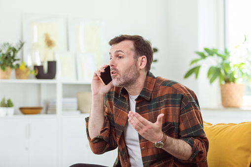 Mature man wearing checkered shirt sitting on sofa at home and talking on smart phone.