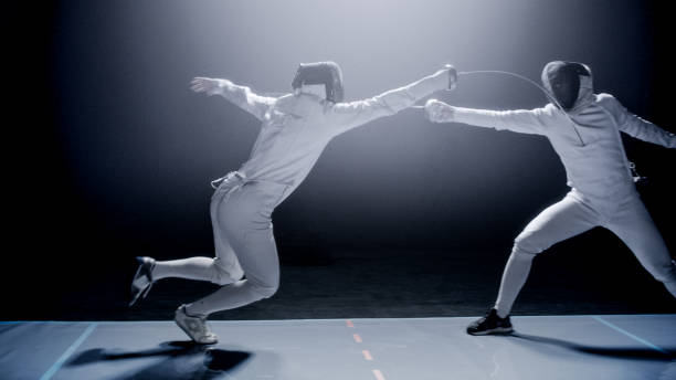 istock Two professional fencers foil swords dueling each other in combat with epic blue stage lighting and smoke effects. Lunging forward 1542472264
