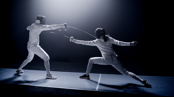 Two fencers dueling in a fencing sports match on lit-up stage. Foil swords clashing to determine the match winner