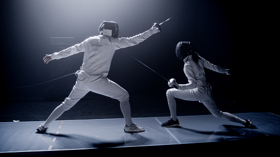 Two fencers dueling in a fencing sports match on lit-up stage. Foil swords clashing to determine the match winner