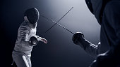 Two professional fencers foil swords facing each other in combat with epic blue stage lighting and smoke effects. Low angle view swords crossed