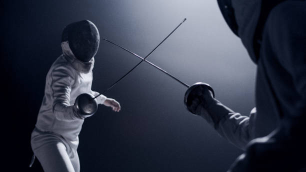 Two professional fencers foil swords facing each other in combat with epic blue stage lighting and smoke effects. Low angle view swords crossed Two fencers dueling in a fencing sports match on lit-up stage. Foil swords clashing to determine the match winner fencing sport stock pictures, royalty-free photos & images