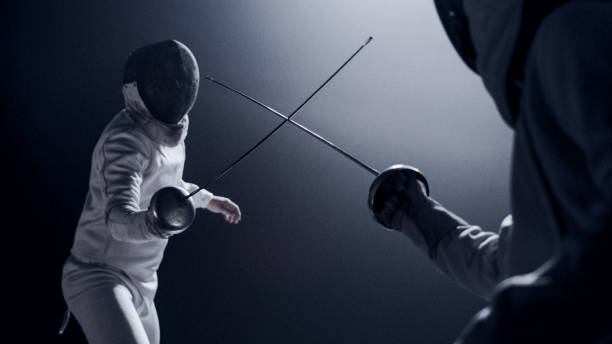 two professional fencers foil swords facing each other in combat with epic blue stage lighting and smoke effects. low angle view swords crossed - fencing sport rivalry sword imagens e fotografias de stock