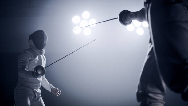istock Two professional fencers foil swords facing each other in combat with epic blue stage lighting and smoke effects. Low angle view challenging 1542473324