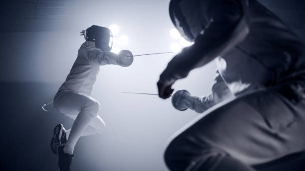 istock Two professional fencers foil swords facing each other in combat with epic blue stage lighting and smoke effects. Low angle view jump 1542473325