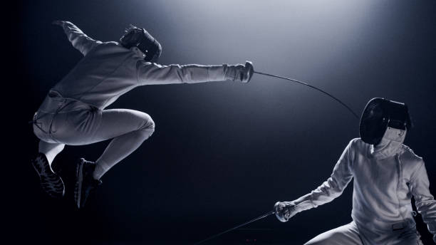istock Two professional fencers foil swords facing each other in combat with epic blue stage lighting and smoke effects. Jumping high up 1542473587