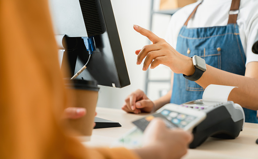 Woman cashier wears an apron and using pos terminal to payment for credit card on coffee shop counter.