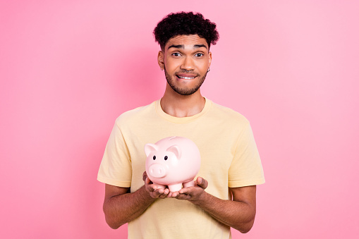 Portrait of young doubtful funny guy wavy hair hold piggy bank collect money unsure about his startup bite lips isolated on pink background.