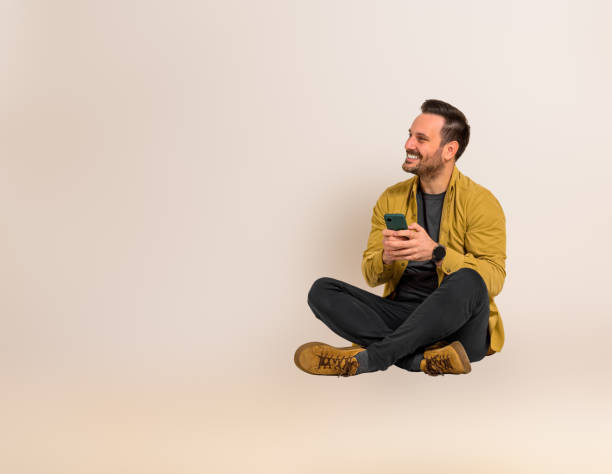 istock Smiling young businessman looking away and text messaging on smart phone while sitting with crossed legs. Handsome professional dressed in casuals doing social media marketing on cellphone against background 1542497426