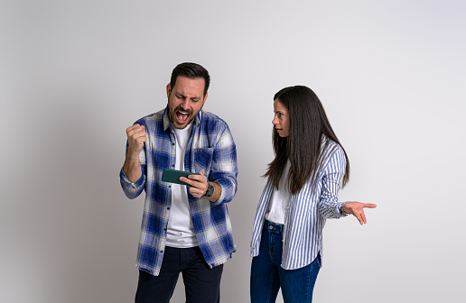 Frustrated girlfriend looking at excited boyfriend playing video game over mobile phone. Irritated young woman standing with male gamer screaming after winning against background