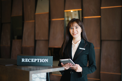 Portrait of happy Chinese female hotel receptionist standing at workplace. Smiling woman receptionist working in hotel.