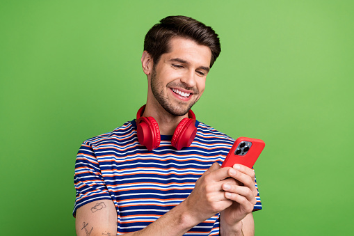 Portrait of cheerful optimistic guy with bristle wear striped t-shirt look at smartphone music app isolated on green color background.