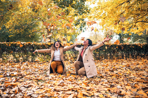 Two smiling young women friends throwing leaves in the park full of fall colors. They are laughing and having fun