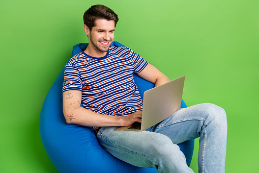 Photo of funky cheerful man dressed striped t-shirt sitting bean bag working modern device isolated green color background.