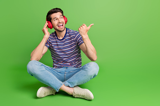 Full length photo of cool excited man dressed striped t-shirt enjoying music looking thumb empty space isolated green color background.
