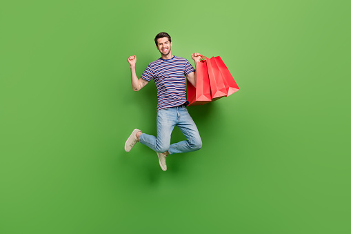 Full length photo of funky lucky man dressed striped t-shirt jumping high rising bargains isolated green color background.