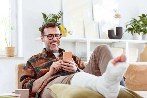 Mature man with broken leg at home Pleased mature man wearing checkered shirt sitting with broken leg on sofa in living room at home and using smart phone. broken leg stock pictures, royalty-free photos & images