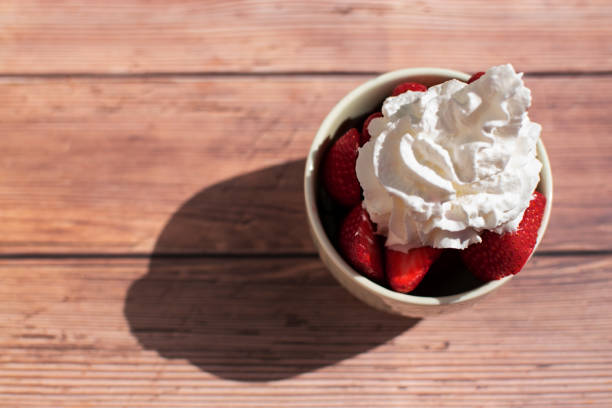 Strawberries cut into pieces covered by whipped cream in ceramic bowl or bowl on rustic outdoor wooden background with sunlight and long shadow, empty space, negative space. Dessert concept,dessert with cream,strawberries with cream,delicious dessert,summer dessert,snack with strawberries,dessert with fruit and cream.View from above, overhead view, view from above.