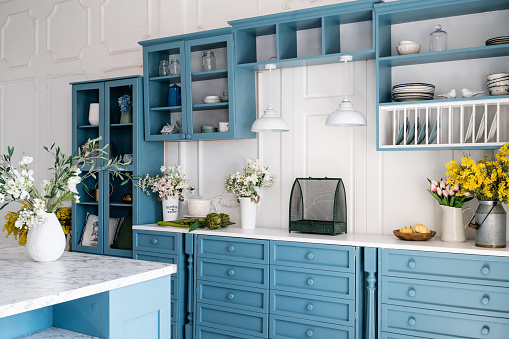 Kitchen with blue vintage furniture, wooden cupboard, kitchenware, drawers, shelves and countertop with jugs and flowers. Home decor against white walls. Monochrome interior.