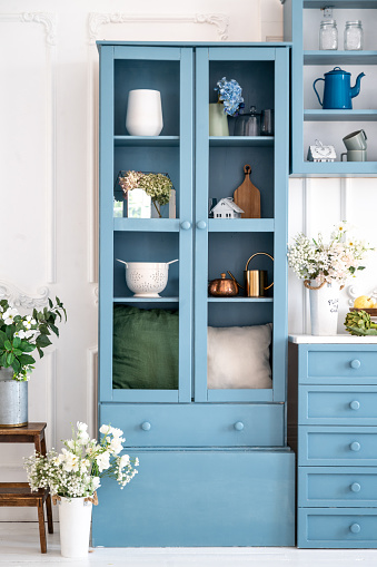 Monochrome blue kitchen. Vertical shot of wooden cupboards with decoration elements. Fresh flowers in vases close to home decor, teapots and jars. Vintage provence style in home interior