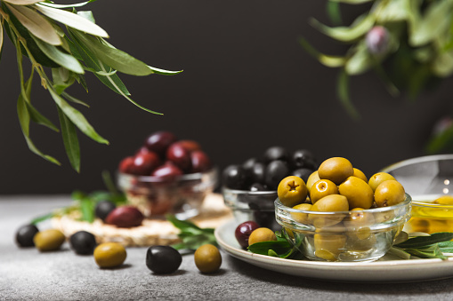 Green, black and red olives on a textured background. Various types of olives in bowls and olive oil with fresh olive leaves. Copy space. Place for text. Mediterranean food. Vegan.