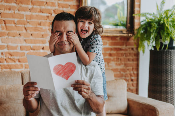 Smiling child hiding father's eyes and handing gift postcard
