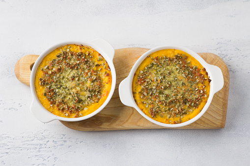 Homemade pumpkin casserole or gratin with cheese and pumpkin seeds in two ceramic tins on a light gray background, flat lay