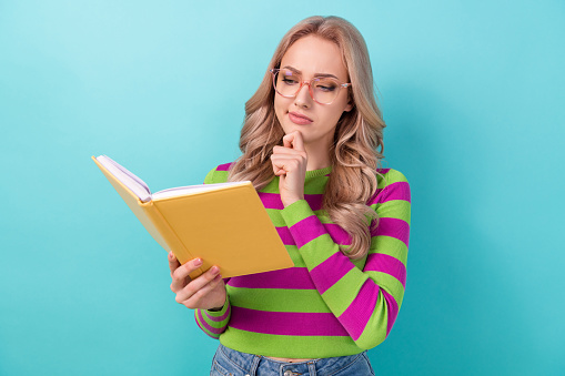 Photo of intelligent minded woman dressed striped sweatshirt look at interesting book hand on chin isolated on teal color background.