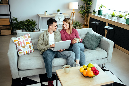 Couple using a digital tablet. They are sitting on a sofa with a modern kitchen behind them. They are attractive, smiling and happy.