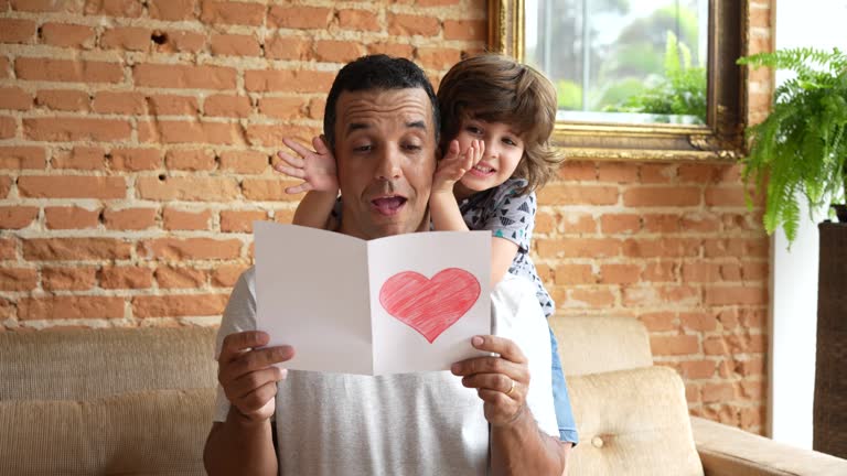 Smiling child hiding father's eyes and handing gift postcard