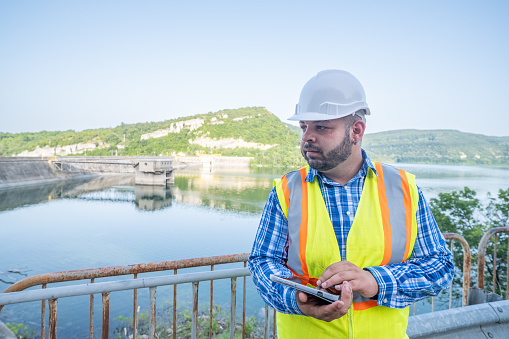 A man working on the maintenance of a dam. He is wearing a blue plaid shirt and a yellow reflective vest on top. He has a white safety helmet and a tablet in his hands.