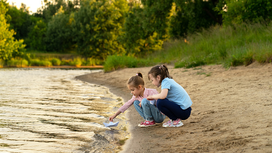 Happy girls fun play with paper boats on river on background of green summer park at sunset. Children walk outdoors