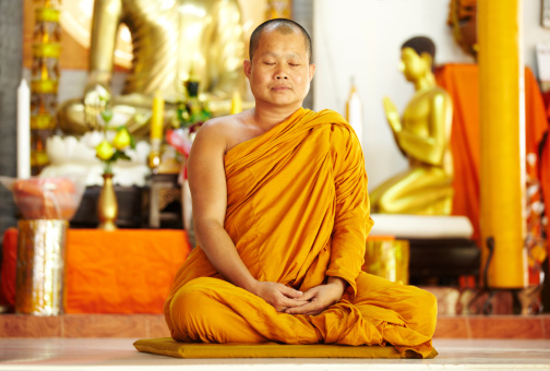 A monk meditating in the lotus position in front of golden Buddhas