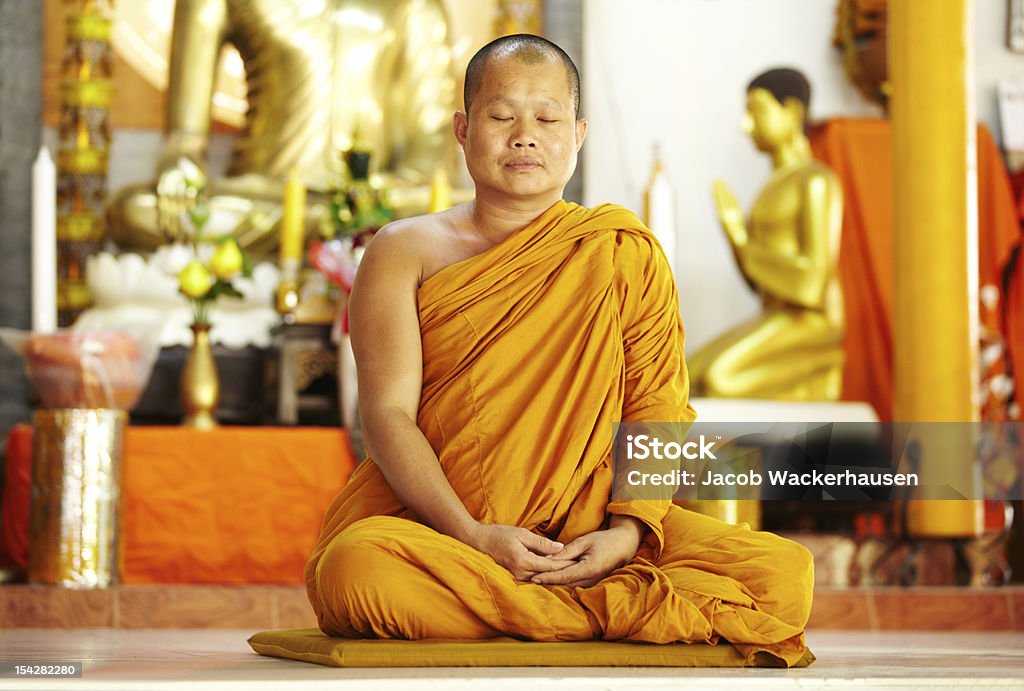 Meditating monk in a sacred shrine A monk meditating in the lotus position in front of golden Buddhas Monk - Religious Occupation Stock Photo