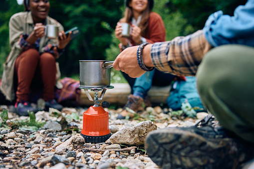 Close up of hiker preparing a drink on portable gas burner while camping with friends in the forest.