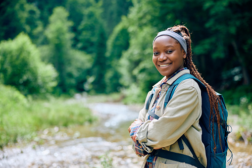 Happy African American woman with backpack hiking in nature and looking at camera.