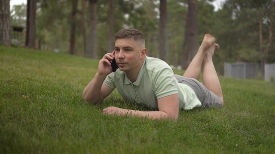 A young man lies on the grass and talks on the phone. A student in the park rests on the grass with communicates by cellular connection. 4k