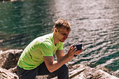 Male portrait of man, who traveling in mountains and taking picture on mobile phone near water. Person doing hiking with sportswear outdoors.