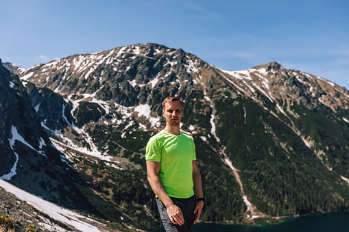 Male portrait of man, who traveling in mountains. Person doing hiking with green sportswear outdoors. Lifestyle photography of real people, tourism concept with smart watch.