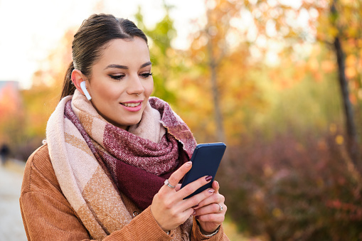 Caucasian young woman using the phone wearing earphones smiling in autumn.