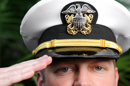 This is a horizontal, color photograph of a United States Navy officer. He is wearing a hat and has his hand up in a salute. Focus on hat.