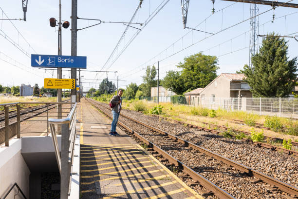 A young man standing on a train platform, waiting to catch a train while on holiday in Toulouse, France. He is looking down the railway track for his train.