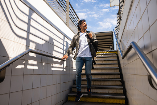 A young man walking down stairs at a train station after arriving on a train while on holiday in Toulouse, France. He is looking to his side and smiling in the sun as he walks down the stairs.